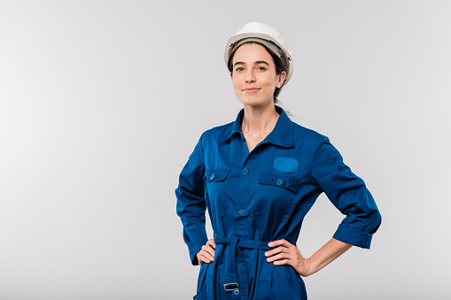 Pretty young successful female engineer in blue workwear and safety helmet keeping her hands on waist in isolation