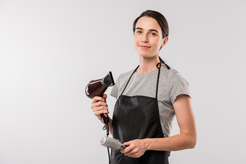 Young professional hairdresser in workwear holding hairbrush and hairdryer for making hairstyle while standing in isolation