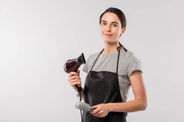 Young professional hairdresser in workwear holding hairbrush and hairdryer Young professional hairdresser in workwear holding hairbrush and hairdryer for making hairstyle while standing in isolation hairdresser stock pictures, royalty-free photos & images