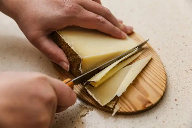 Woman's hand holds a yellow-brown cured sheep cheese and brown crust and with the other hand she has a special knife with holes and wooden handle to cut the cheese on a pine cutting board that is located on top of marble Clear kitchen there are also some cheese triangle cuts
