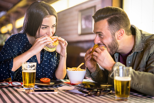 Happy couple having fun eating burger at restaurant pub fast food - Young people enjoying moment at indoor diner location - Relationship concept with focus on girl face with warm vivid filter