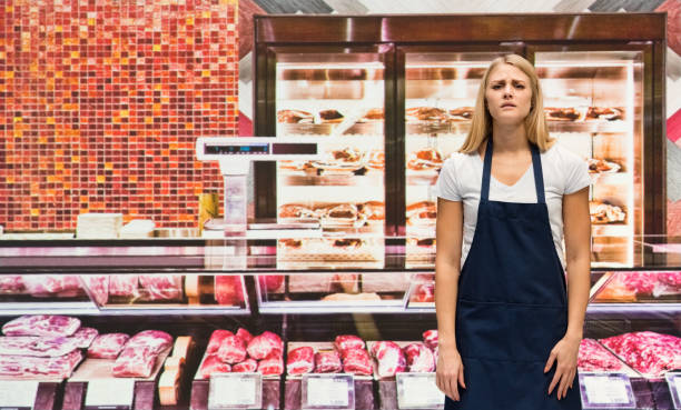 One person / waist up of 20-29 years old adult beautiful blond hair caucasian female / young women owner / butcher standing at the supermarket / delicatessen / butcher's shop / market - retail space / refrigerated section in the store wearing t-shirt / shirt / apron who is feeling sad / depression - sadness / emotional stress / with anxiety / sulking / selling / meat / lamb - meat / meat chop / brick wall / retail / food una persona / cintura para arriba de 20-29 años de edad adulto hermoso cabello rubio mujer caucásica / joven mujeres dueña / carnicero de pie en el supermercado / delicatessen / carnicería / mercado - espacio de venta al por menor / sección refrigerad