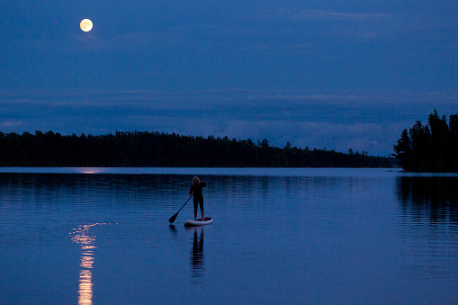 A woman goes for a stand up paddleboard trip at night under a full moon on a small lake in Ontario, Canada.