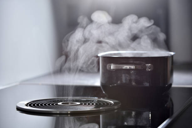 Modern electric induction cooker with built-in ventilation and extractor hood which draws steam from boiling water in a pan. Steam from a boiling pot is drawn into the integrated range hood stock photo