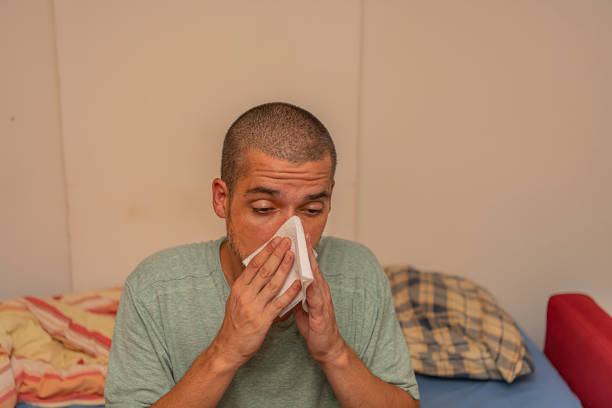 Closeup Portrait of Mature Ill Man with Allergy or Cold, Blowing his Nose with a Tissue, Looking Miserable Unwell and Very Sick. Man is Sitting and Home With Allergy Problems and Blowing a Nose with Tissue Papers. Flu Season, Vaccination.
