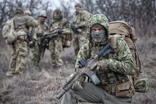 Shoulder portrait of army infantry soldier, tactical group member in camouflage uniform, carrying backpack, wearing camo jacket hood, hiding face with mask, looking in camera, comrades on background