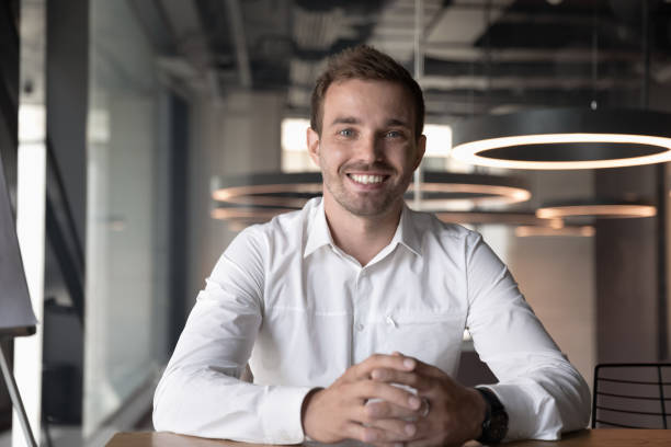 Portrait of smiling male employee posing in office Portrait of smiling male Caucasian employee sit at office desk look at camera in modern office, happy man worker millennial businessman posing, having video call with client or customer online head shoot stock pictures, royalty-free photos & images
