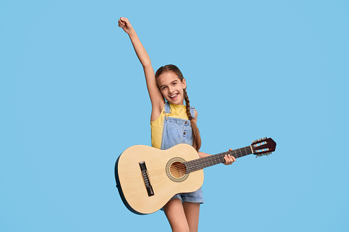 Excited little girl raising arms and smiling while playing acoustic guitar against blue background