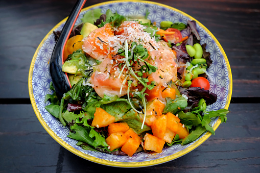 Close up shot of healthy salad bowl with sliced raw salmon ,avocado, mango and leaf vegetables above dark wooden table