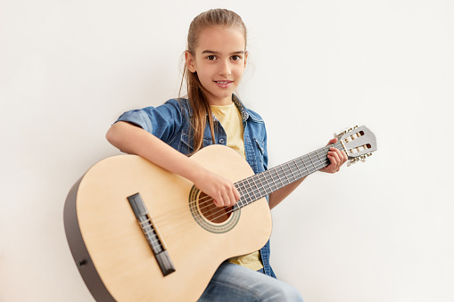 Glad kid in casual outfit looking at camera and learning to play guitar against white wall at home