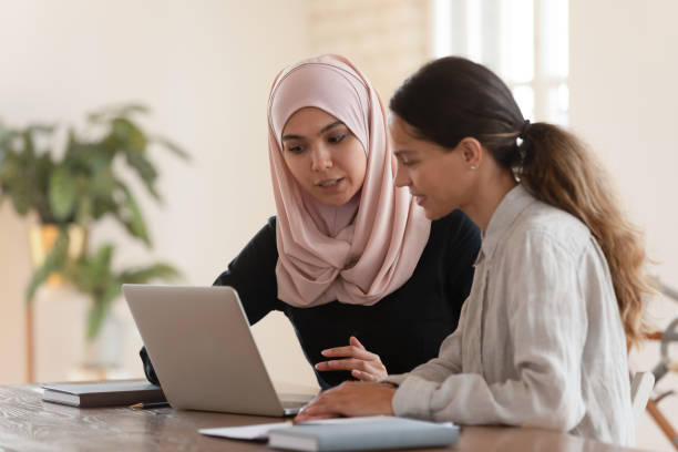 Focused team leader training millennial female intern. Concentrated young arabian woman in hijab sitting with smiling colleague at table, looking at computer screen, explaining new company software. Focused team leader training millennial female intern. arab woman stock pictures, royalty-free photos & images