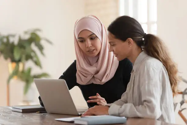 Concentrated young arabian woman in hijab sitting with smiling colleague at table, looking at computer screen, explaining new company software. Focused team leader training millennial female intern.