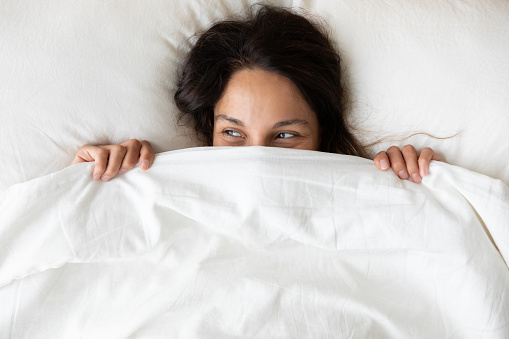 Above close up view young millennial cheerful woman lying under blanket inside of cozy bed on white soft pillow hiding her face, pretty curious 30s female feeling shy peeking looking away from duvet