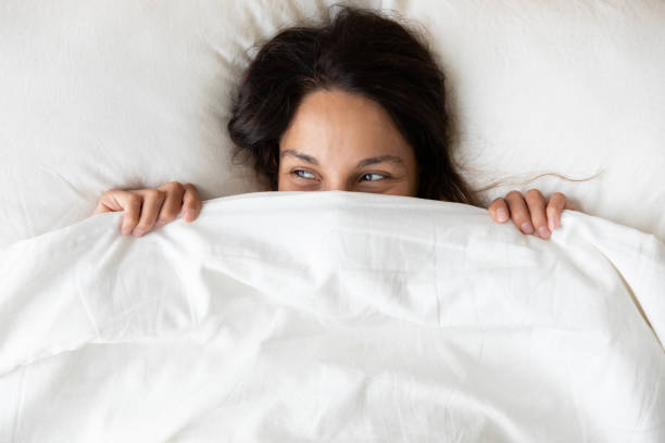 Above view playful young woman hiding face under blanket Above close up view young millennial cheerful woman lying under blanket inside of cozy bed on white soft pillow hiding her face, pretty curious 30s female feeling shy peeking looking away from duvet duvet stock pictures, royalty-free photos & images