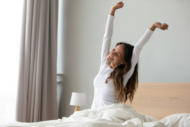 Woman sitting in bed stretching her arms muscles after sleep Happy woman in white nightwear sitting in bed awakened from enough and healthy sleep feels good, stretching her arms muscles after sleep and long immobility wakes up start new day with smile concept waking up photos stock pictures, royalty-free photos & images
