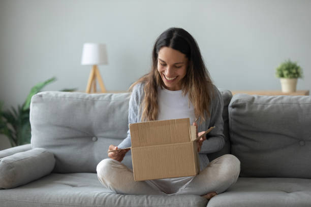 Overjoyed young woman opens box parcel feels satisfied Overjoyed woman seated on sofa hold on lap small cardboard box open parcel client feels satisfied bought goods in internet, happy addressee received package from friend, quick delivery service concept receiving box stock pictures, royalty-free photos & images