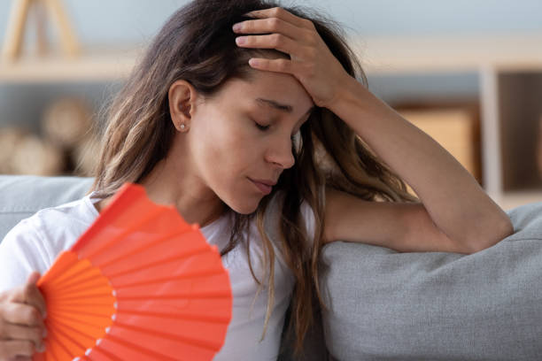Close up woman sitting on couch reducing heat using fan Close up view millennial woman sitting on couch exhausted by hot weather closed eyes feels unwell dying of heat holding in hand orange fan using it for reducing too hot temperature indoors concept hormone stock pictures, royalty-free photos & images