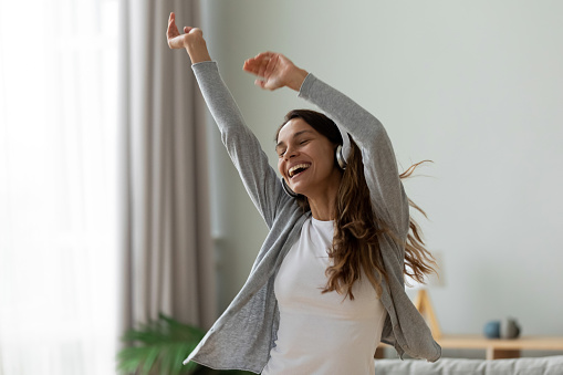 In cozy living room carefree happy woman raised hands dancing wearing headphones listening favourite song music through wireless modern device, enjoy life feels overjoyed, concept of pastime and hobby