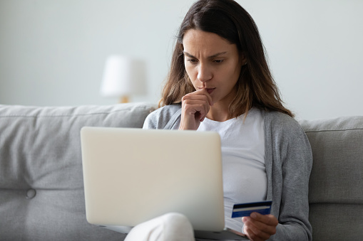Concerned woman sit on sofa holding on lap pc in hand credit card feels puzzled about money lack, forgot password, problem with purchasing through internet, no access to online e-bank services concept