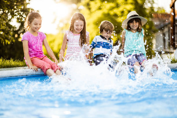 Adorable toddler girl ducks away from splashing water fountain while walking hand in hand with her older sister through a water park. The little girl's older cousins and grandmother are in the background. The splash pad is in a public park surrounded by fir trees in the Pacific Northwest.
