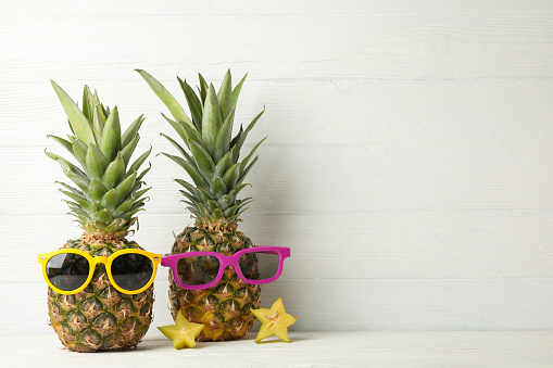 Pineapples with sunglasses on white wooden background, space for text