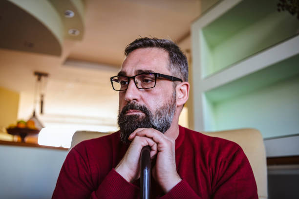Depressed Mature Man with Eyeglasses and Beard is Feeling Sad and Thoughtful. Man with Problems is Sitting in Armchair at Home and Holding a Walking Cane and is Looking Away.