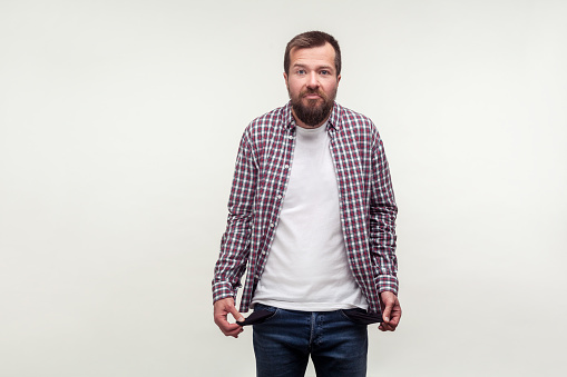 Portrait of bankrupt bearded man in casual plaid shirt standing upset with depressed face expression, turning out empty pockets has no money, low salary. studio shot isolated on white background