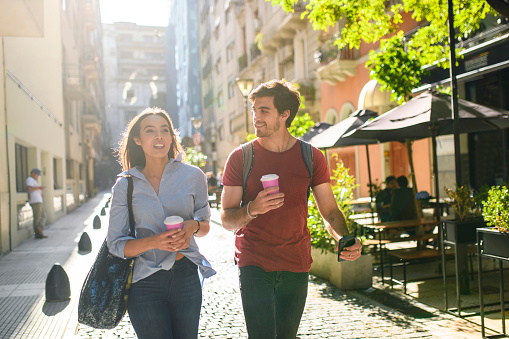 Hispanic male and female tourists in their 20s walking with coffee and smart phone down cobblestone side street in Buenos Aires.