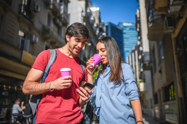 Low angle view of young Hispanic male and female travelers enjoying coffee and checking smart phone for directions in Buenos Aires.
