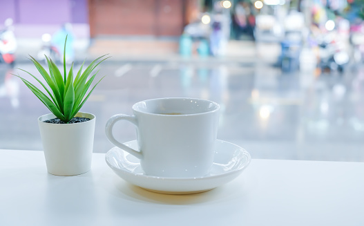 A cup of coffee on white table in modern coffee shop on a rainy day, close-up shot of a white ceramic cup of coffee and cactus on white table with a view of city street blurred background.