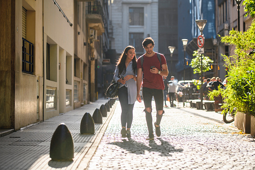 Hispanic couple in their 20s walking down cobblestone side street in Buenos Aires and checking smart phone.