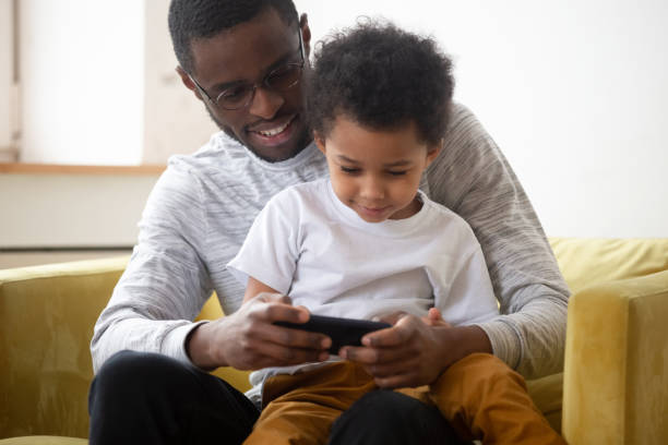 o pai americano africano novo joga no telemóvel com filho pequeno - digital tablet family father offspring - fotografias e filmes do acervo