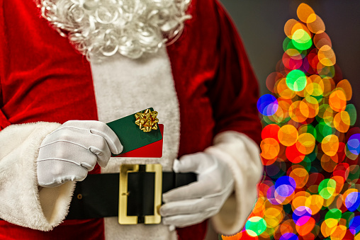 Santa Claus holding a pair of gift cards with a gold bow and in the background the bokeh of multi-colored Christmas tree lights