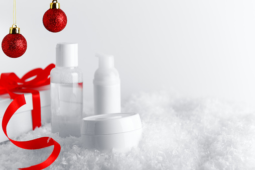 Skincare cosmetics bottles on stylized snow covered surface. White gift box with red ribbon and glittery baubles decorative background. New Year and Christmas time woman presents idea.