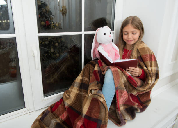 Imagination. Little child read book to bunny toy on Christmas eve. Little girl enjoy reading Christmas story. Little reader wrapped in plaid sit on window sill. Childrens book. Magic xmas spirit. imaginación. el niño leyó el libro para el juguete de conejo en la víspera de navidad. niña disfruta leyendo la historia de navidad. pequeño lector envuelto en a cuadros sentarse en el alféizar de la ventana. libro para niños. espíritu mágico de