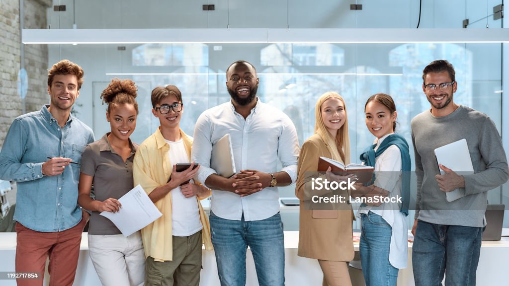 Dream work. Portrait of young and successful co-workers in casual wear smiling at camera while standing in working space Portrait of young and successful co-workers in casual wear smiling at camera while standing in working space. Teamwork concept. Collaboration Multiracial Group Stock Photo
