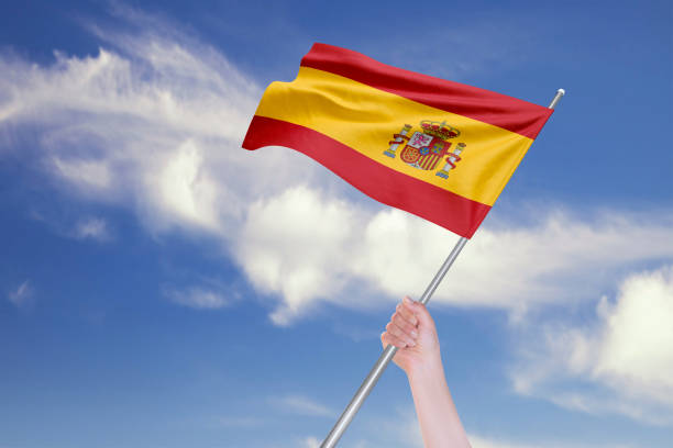 istock Female Hand is Waving Spanish Flag Against Blue Sky with Clouds 1192720528