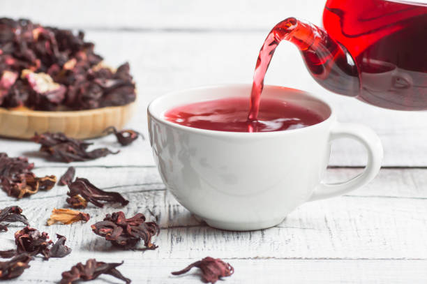 White cup of healthy hibiscus tea pouring from the teapot with dried hibiscus flowers on white wooden background, winter hot drink concept for cold and flu stock photo