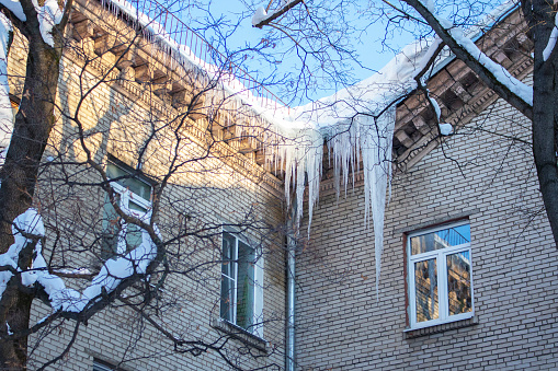 Big long icicles hanging from the roof of a residential building, winter ice cold snow nature. Freezing ice water at the corner of a brick apartment building, danger