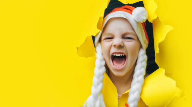크리스마스 선물을 기다리고 있습니다. 빨간 산타 모자를 쓰고 피그테일을 입은 아름다운 작은 소녀가 노란색 파스텔 톤의 구멍을 내다보고 있다. 크리스마스와 새해 개념입니다. 공간을 복사� - human mouth paper screaming hole 뉴스 사진 이미지