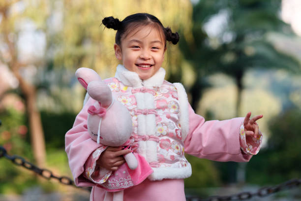 retrato da menina chinesa na roupa do ano novo que sorri na câmera com gesto da vitória e brinquedo do coelho em seu braço, 4-5 anos velho, conceito feliz do ano novo. - child 4 5 years laughing little girls - fotografias e filmes do acervo