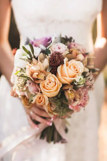 Photo of Bride holding in hands small wedding bouquet in orange autumn colors. Pink and orange roses, white peony, dried flowers and leaves.