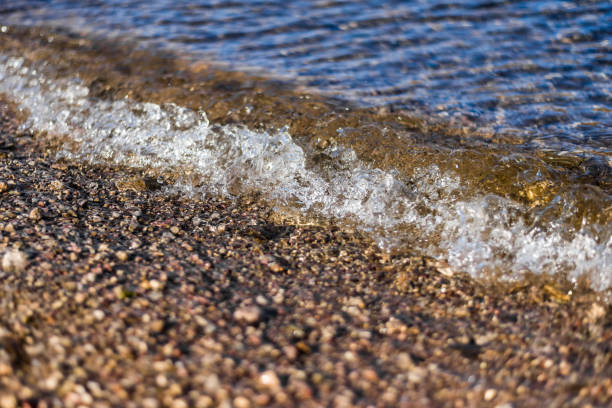 Waves roll gently over a pebbled beach, creating white foam as the sea meets the shore. A tranquil coastal scene with textured rocks and flowing water, suitable for nature and travel themes.