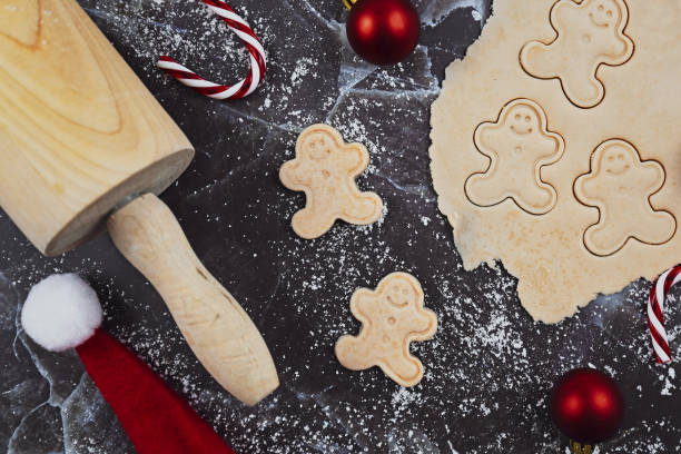 overhead-flach-lay-konzept zum backen von weihnachtsplätzchen mit ausgerolltem keksteig, keksen in form von fröhlichen lebkuchenmännern und saisonaler dekoration wie kugeln auf dunklem marmorhintergrund - candy cane christmas cookie raw food stock-fotos und bilder