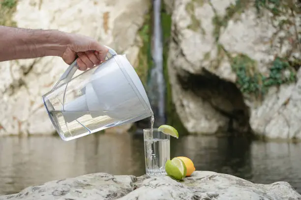 Photo of A hand pouring filtered water in the big glass with lemon on the nature background. Filter jug