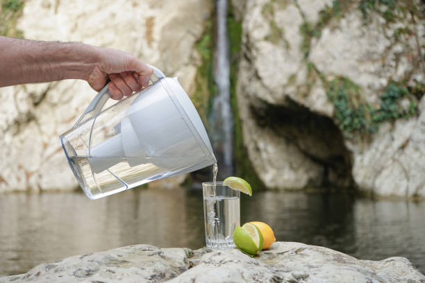 a hand pouring filtered water in the big glass with lemon on the nature background. filter jug - mineral waterfall water flowing imagens e fotografias de stock