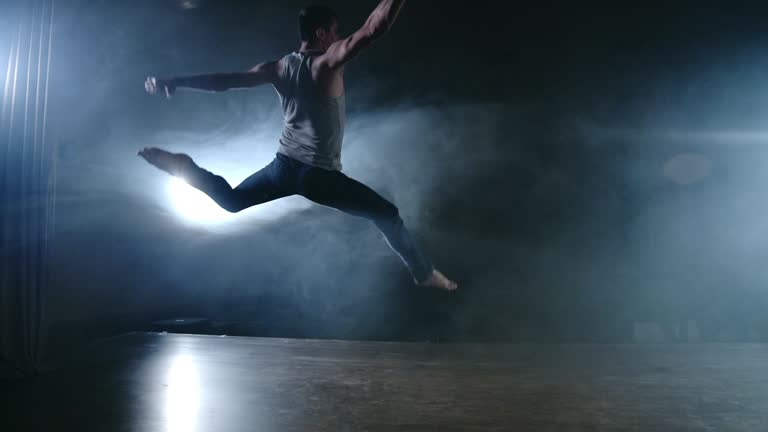 A modern ballet, a man performs jumps and spins in the light of spotlights and smoke on a dark background. Acrobatic choreography, rehearsal of the script of modern ballet