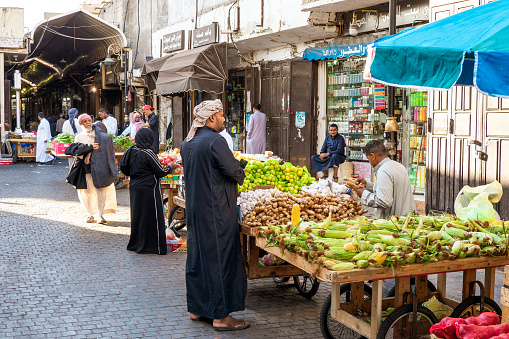 December 7th 2019, Jeddah, Saudi Arabia: The Souq Al-Hababa at the Souk Baab Makkah Street is a busy vegetable and fruit street market in Balad, Jeddah. On this bustling street, vendors advertise their best fruits, potential customers are looking at the products. People strolling along the market. A man in traditional clothing buys a kind of root from a vendor. Apart from the grunge, that's an amazing place!