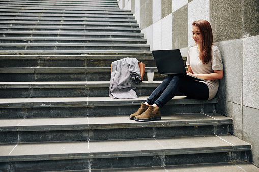 Smiling female freelancer sitting on steps outdoors, working on laptop and writing articles