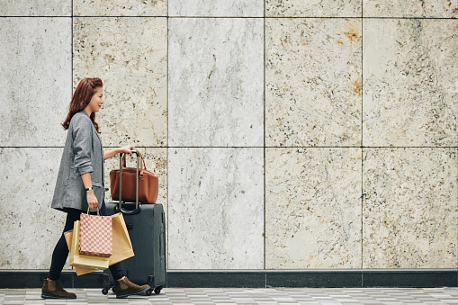 Smiling Vietnamese woman with many shopping bags and suitcase walking along granite wall